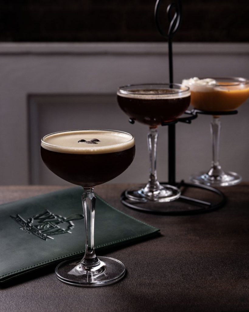 A dark espresso martini garnished with coffee beans sits on a textured table beside a green leather-bound menu, with two additional coffee cocktails elegantly displayed on a black metal stand in the background